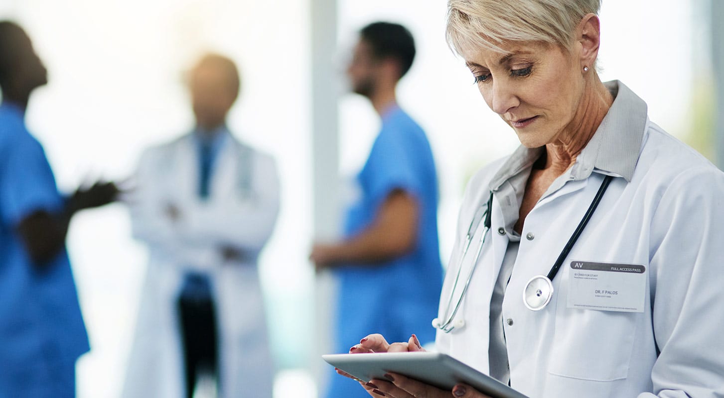 A female doctor in a white coat is holding a tablet, with a team of doctors and nurses conversing in the background.